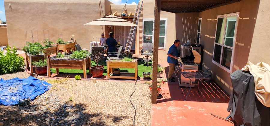 A backyard patio featuring multiple raised garden beds, a grill station, and workers preparing food under a large umbrella, while others work on a nearby ladder.