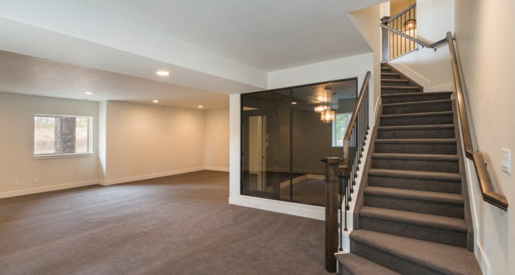 Spacious Basement Living Area Large basement living room with gray carpeting, white walls, a staircase, and a mirrored partition reflecting a dark furnished room.