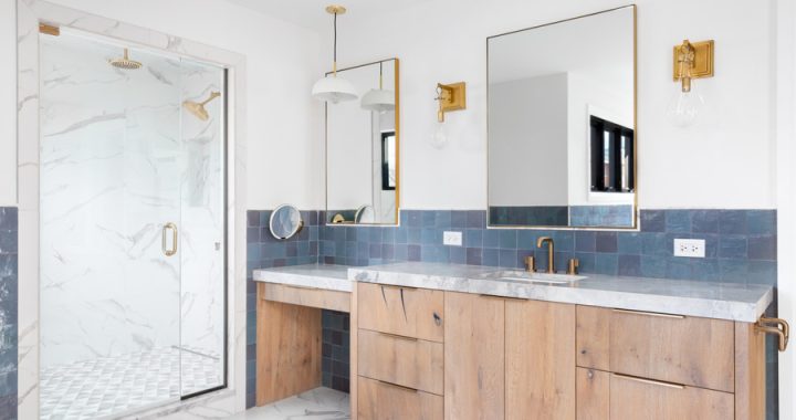 A bathroom with a wood cabinet, gold accents, and a marble shower. A luxurious bathroom with a wood vanity cabinet, blue tiles on the walls, gold accented lights and mirrors, and a large walk-in shower lined with marble tiles.